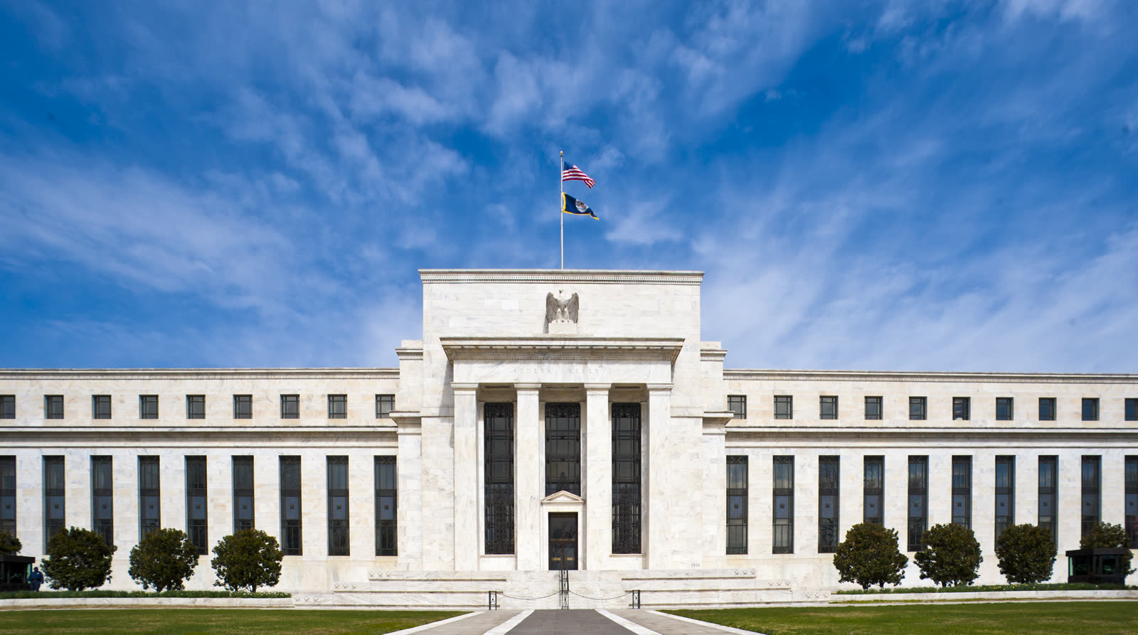 La façade du bâtiment Eccles de la Réserve fédérale à Washington, colonnes de marbre blanc et drapeaux au sommet, sous un ciel bleu